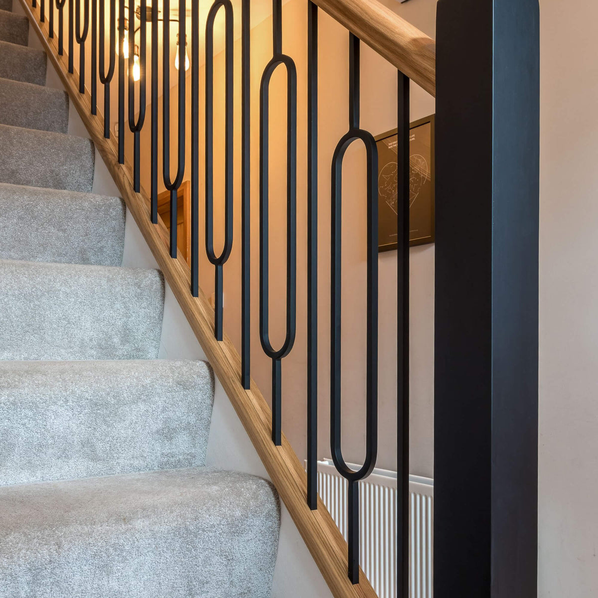 A close-up view of a staircase with light grey carpet, a natural wood handrail, and Black Oval Spindles.