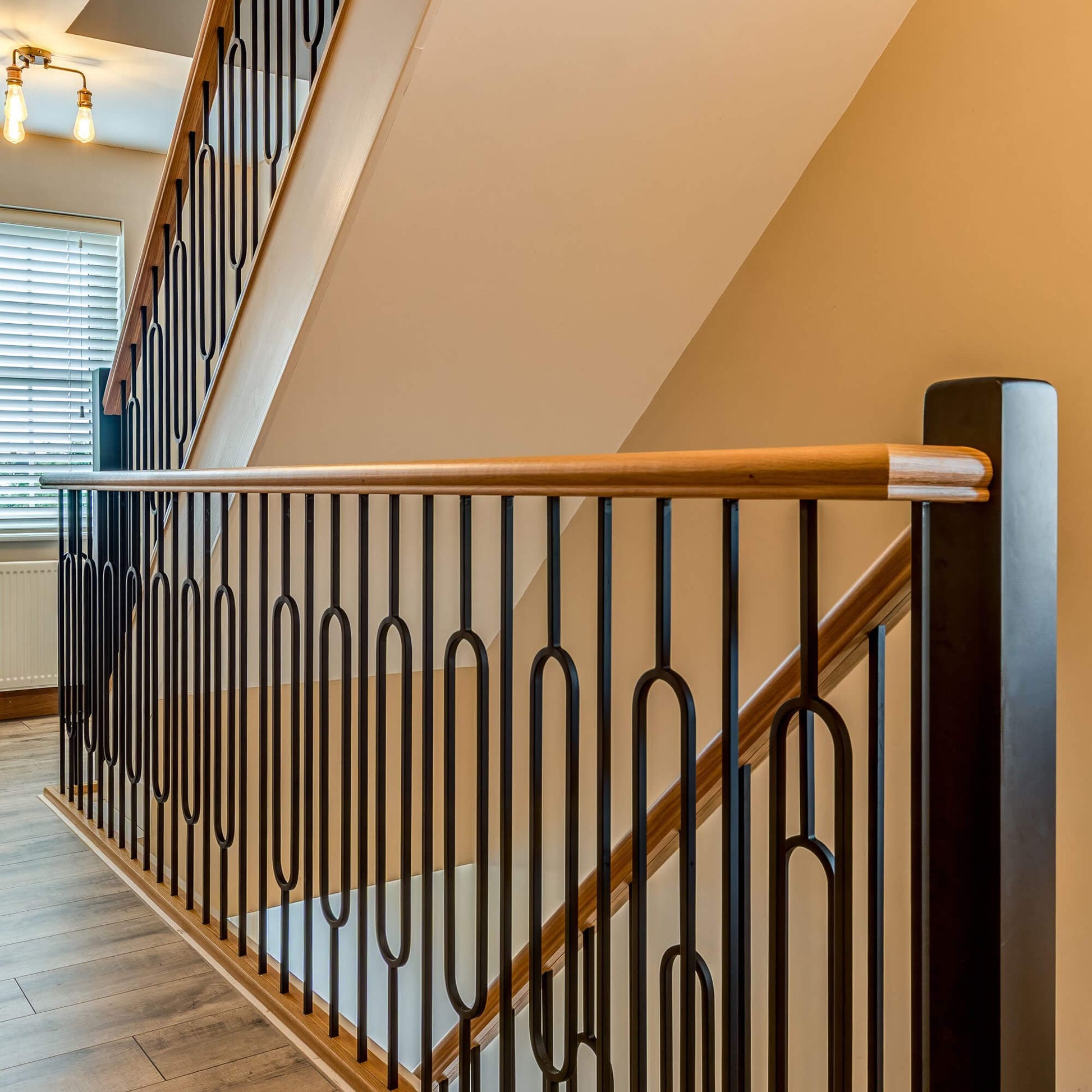 A modern staircase railing with Black Oval Spindles, white oak wood handrail, and a dark wood post, set against a warm beige interior wall.