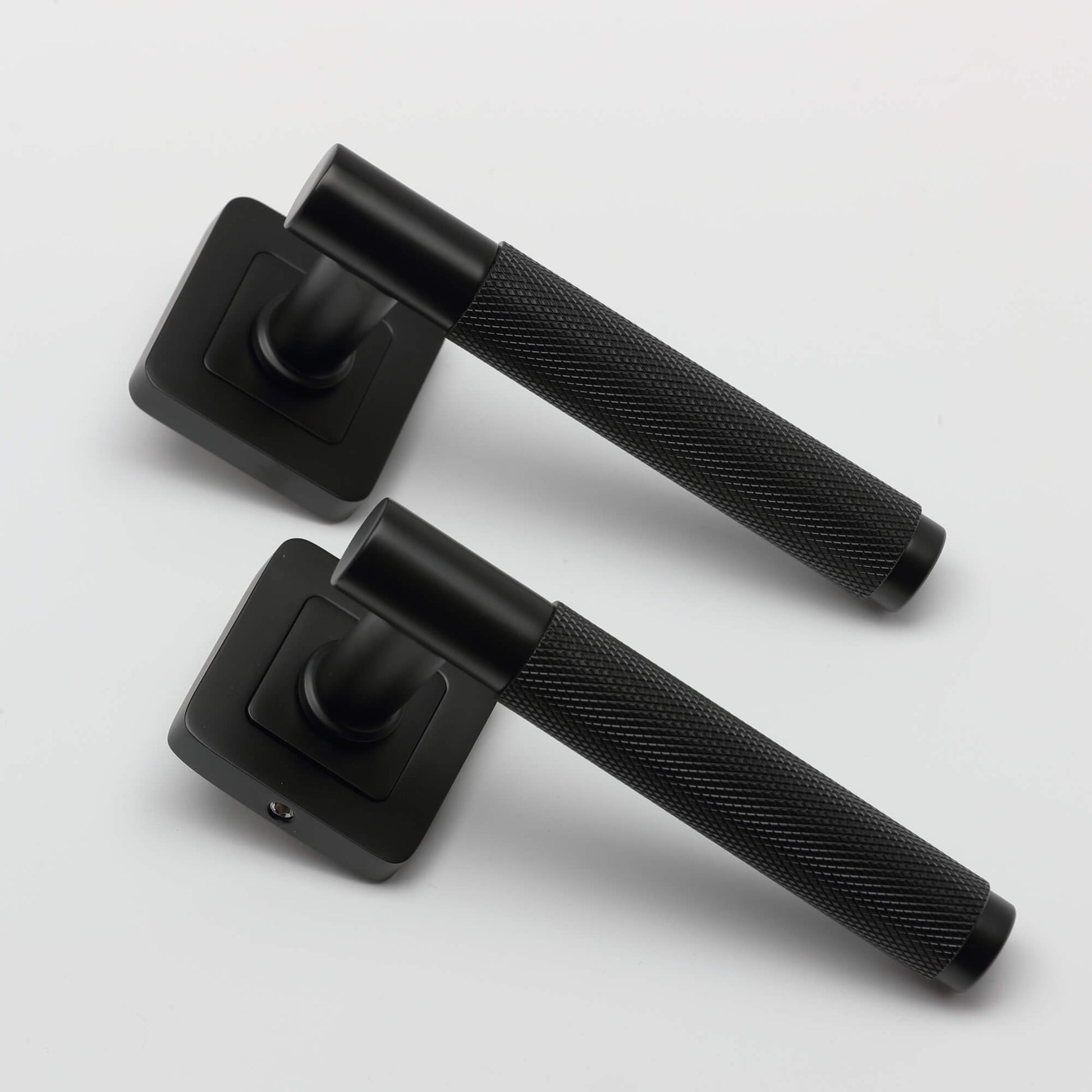 An angled view of a pair of Knurled Flat Black Square Rose door handle on a light grey background.
